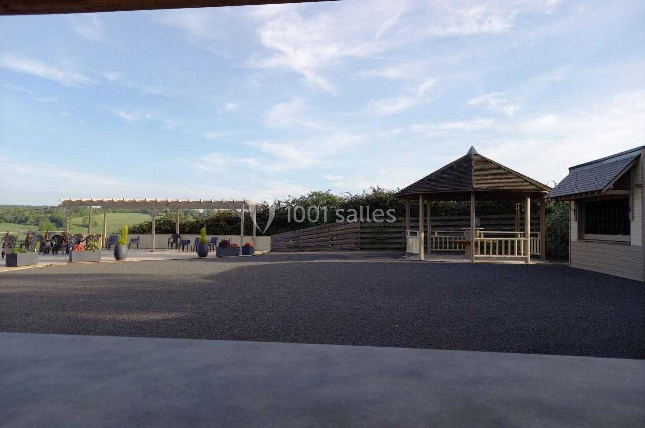 Cour extérieure avec un kiosque en bois, une pergola et des tables, entourée de verdure sous un ciel dégagé.