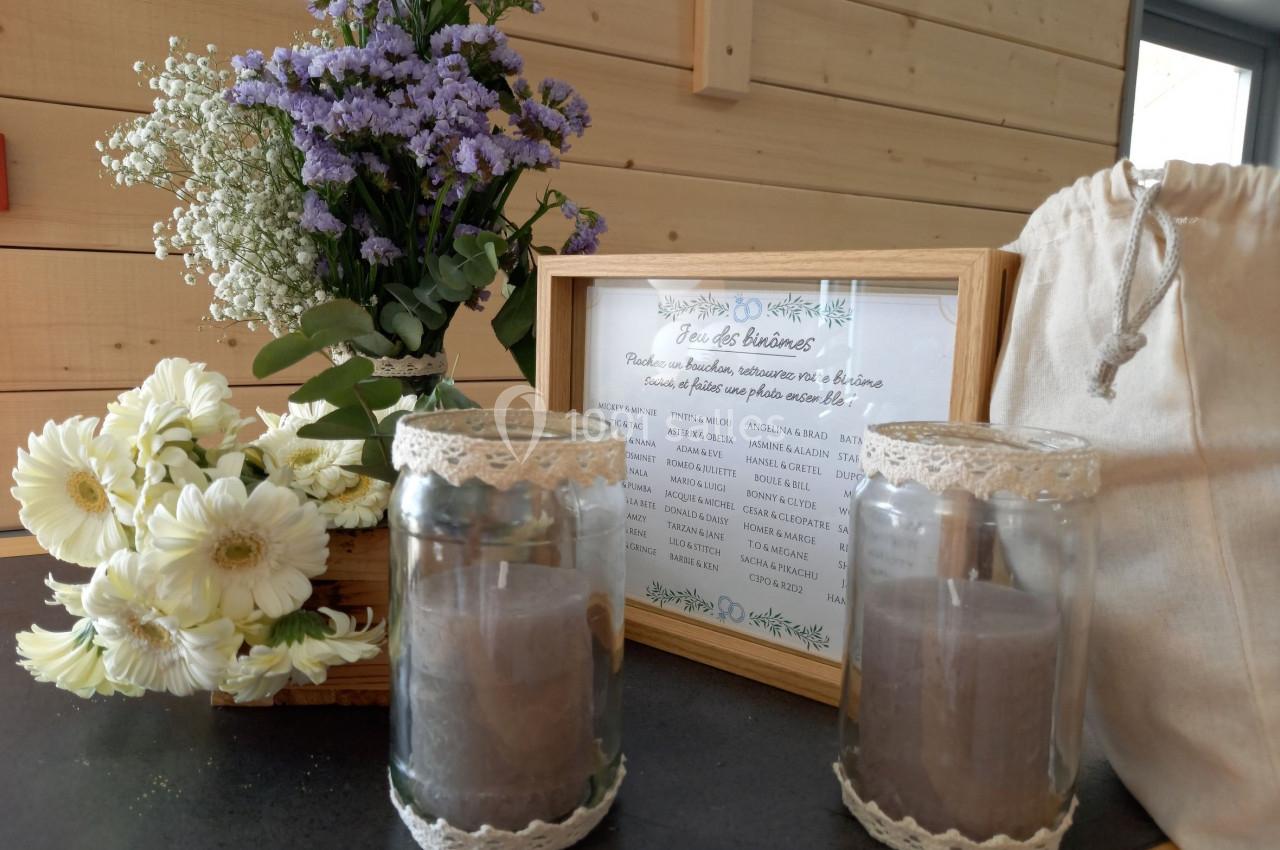 Bougies décoratives en verre, bouquet de fleurs et cadre avec texte sur une table en bois dans un intérieur lumineux.
