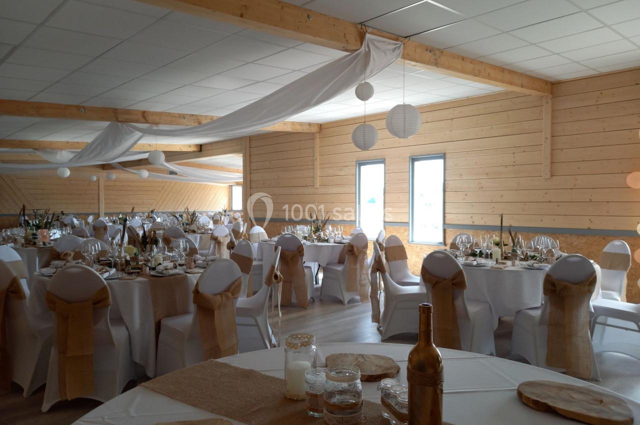 Salle de réception décorée avec des tables rondes, nappes blanches, chaises ornées de nœuds beige et éclairage naturel.