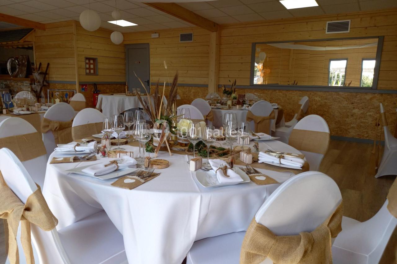 Salle décorée pour un événement avec des tables rondes dressées, nappes blanches et chaises ornées de nœuds en toile de jute.