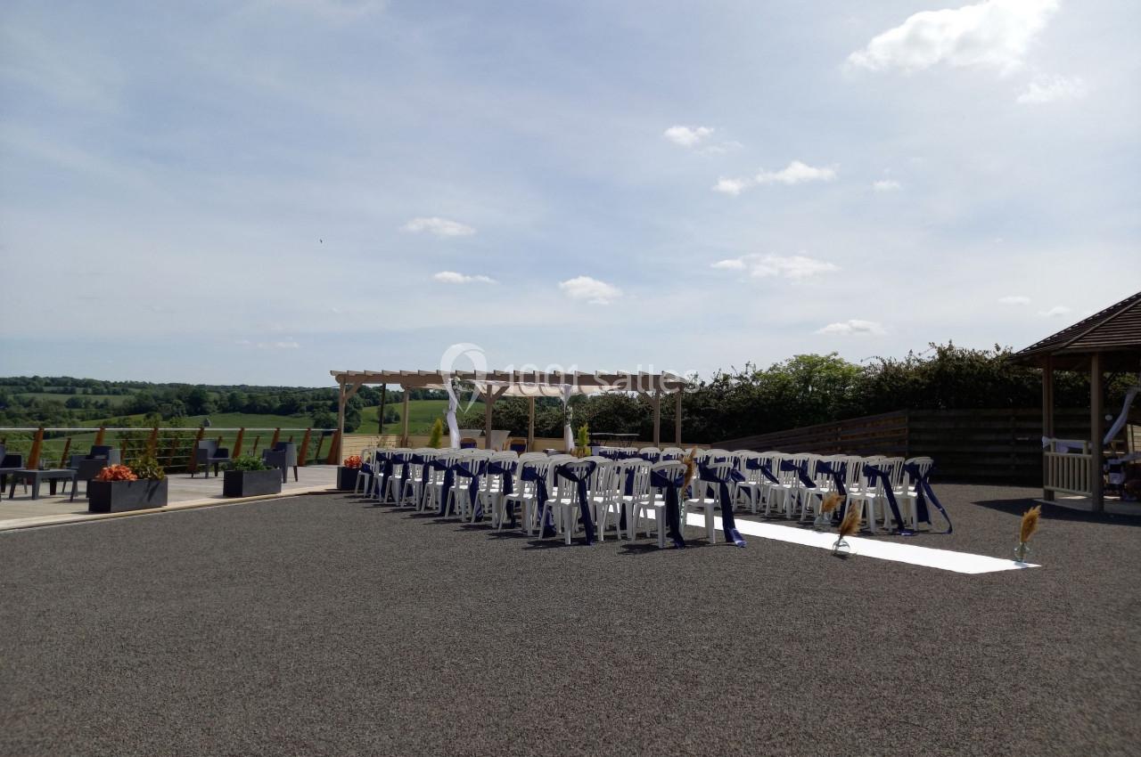 Chaises alignées en extérieur sous une pergola, prêtes pour une cérémonie, avec vue sur un paysage champêtre.