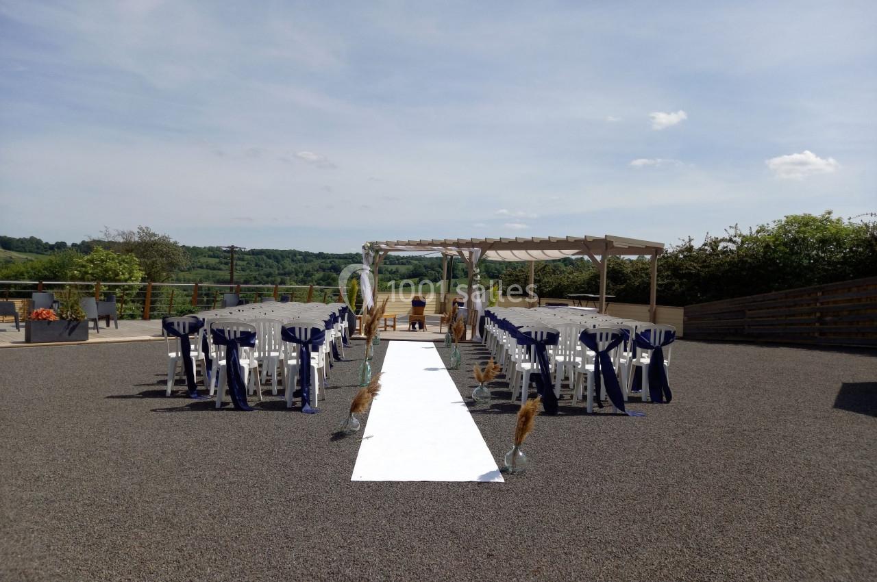 Allée centrale blanche bordée de chaises décorées en extérieur, menant à une pergola avec vue sur la campagne.