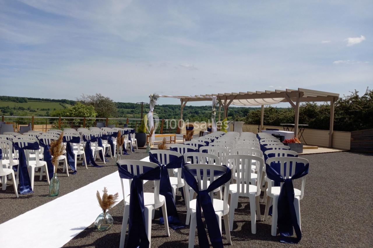 Chaises blanches alignées avec rubans bleus, allée centrale et pergola en extérieur sous un ciel dégagé.