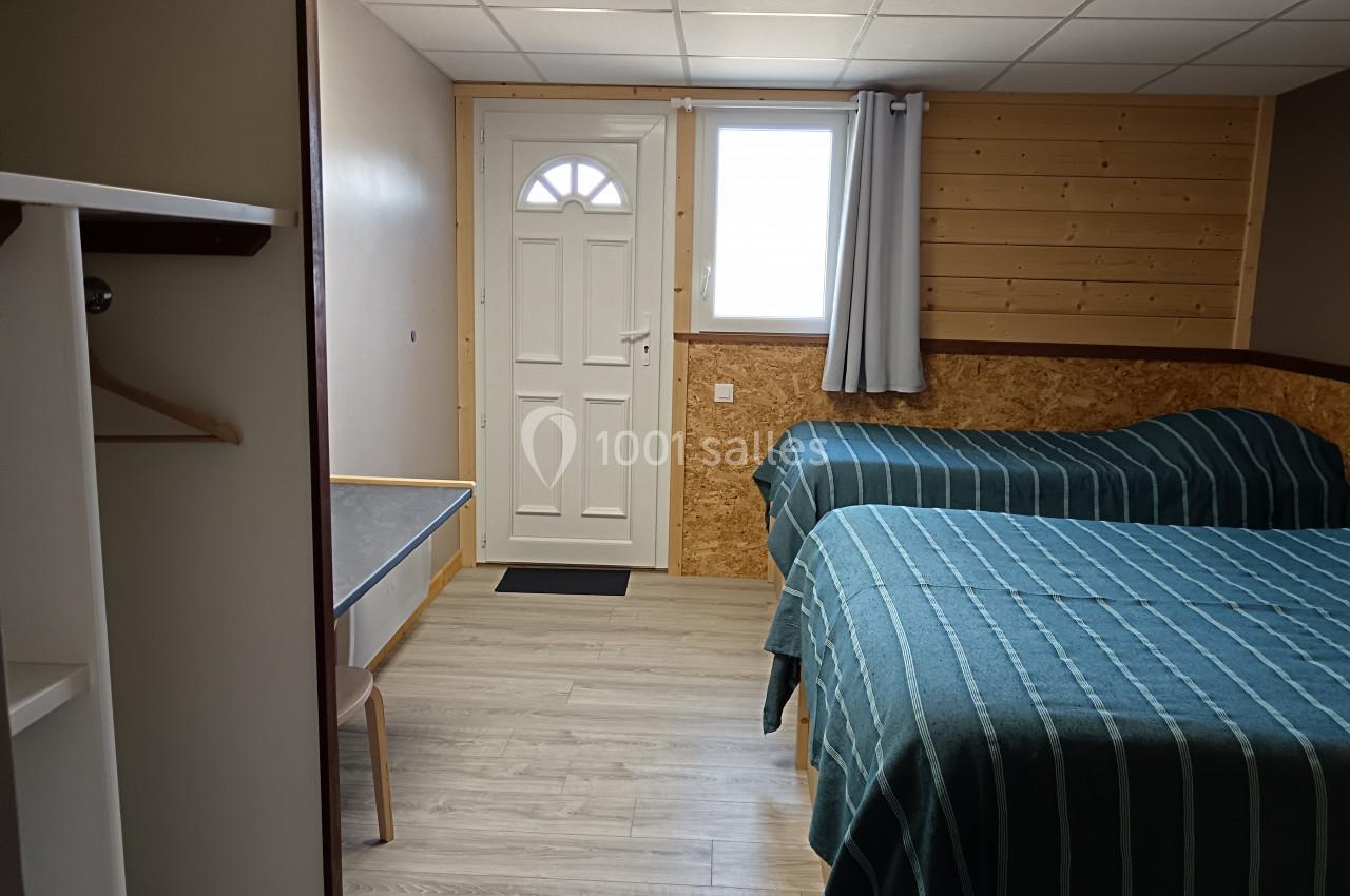 Chambre simple avec deux lits, murs en bois, sol clair, une table, une porte et une fenêtre avec rideau gris.