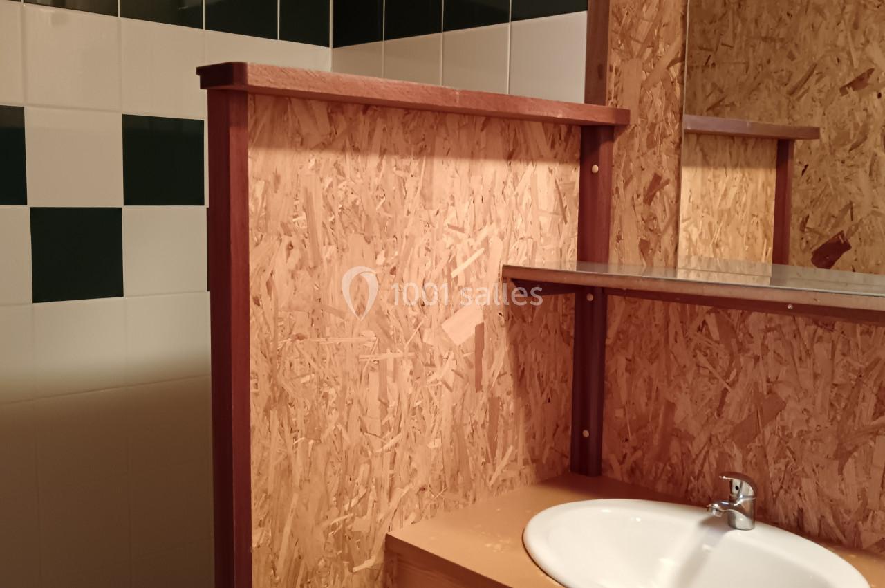 Lavabo avec plan de travail en bois, miroir et cloison en panneaux OSB, dans une salle de bain partiellement carrelée.