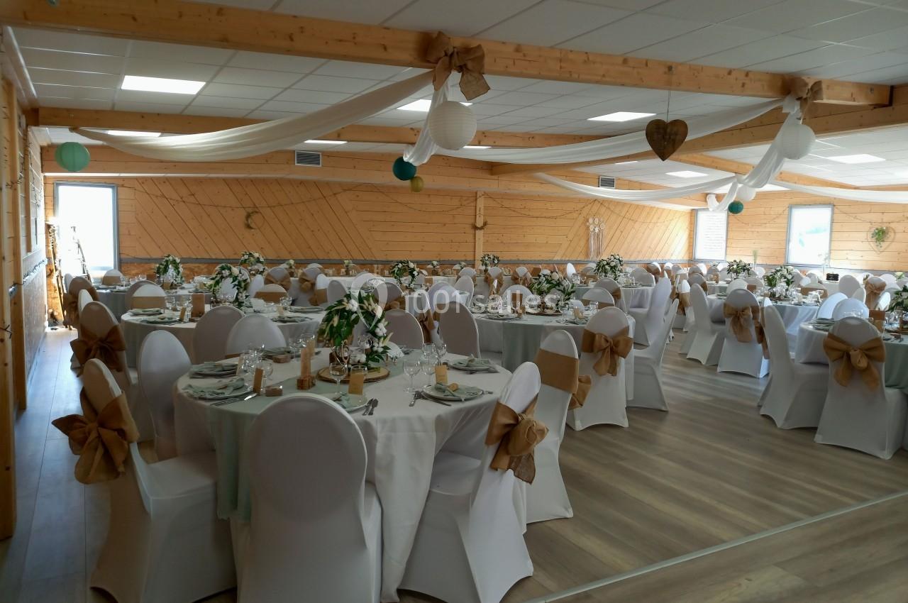 Salle de réception décorée avec des tables rondes, nappes blanches, chaises ornées de nœuds beiges et guirlandes suspendues.