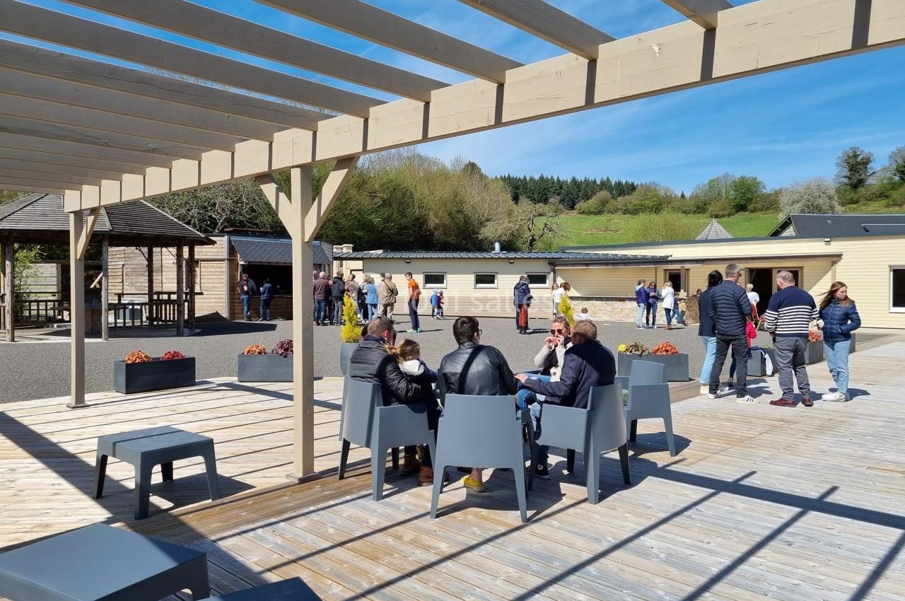 Des personnes assises et debout sur une terrasse en bois, sous une pergola, avec des bâtiments et un paysage en arrière-plan.