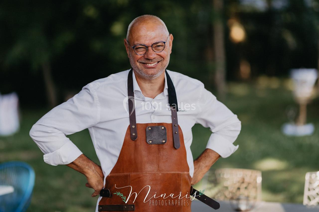 Homme souriant portant un tablier en cuir, debout en extérieur avec un fond flou de verdure et de mobilier.