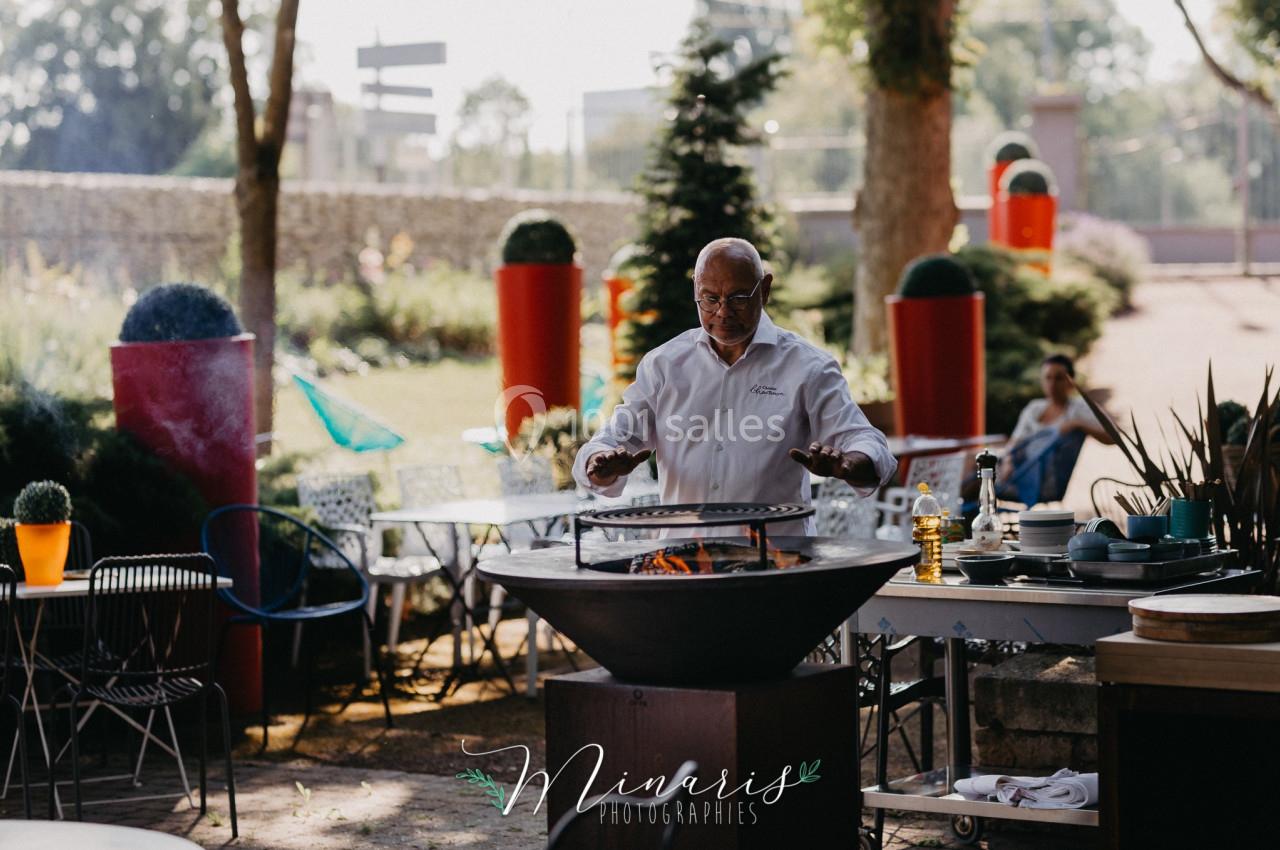 Un chef cuisine sur un barbecue rond dans un jardin aménagé avec des tables et des plantes.