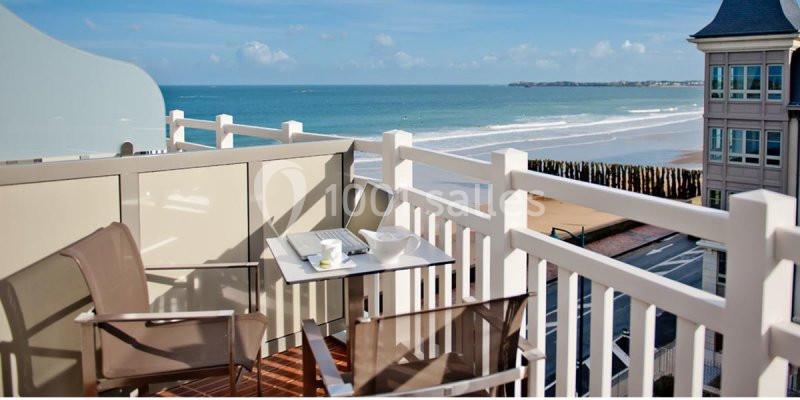 Vue depuis un balcon avec table et chaises, donnant sur une plage, l'océan et des bâtiments en bord de mer.