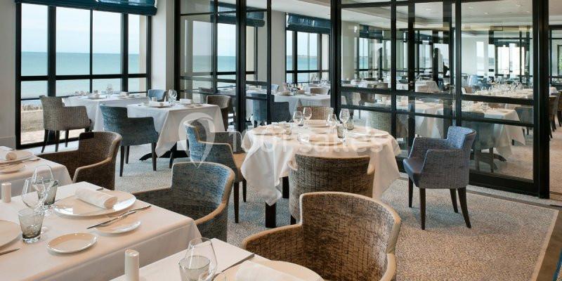 Salle de restaurant élégante avec des tables dressées, des chaises modernes et une vue sur la mer à travers de grandes baies…