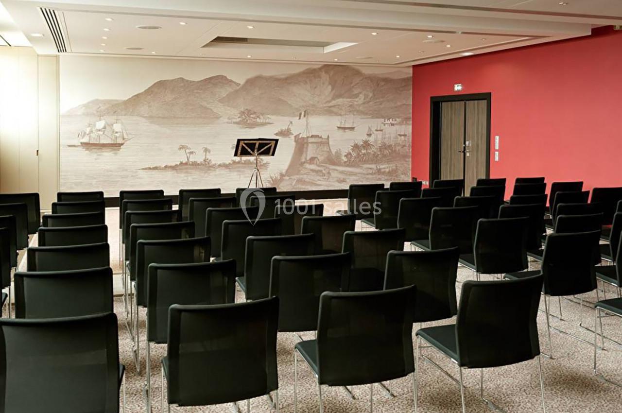 Salle de conférence vide avec des rangées de chaises noires, un mur rouge et une fresque représentant un paysage maritime.