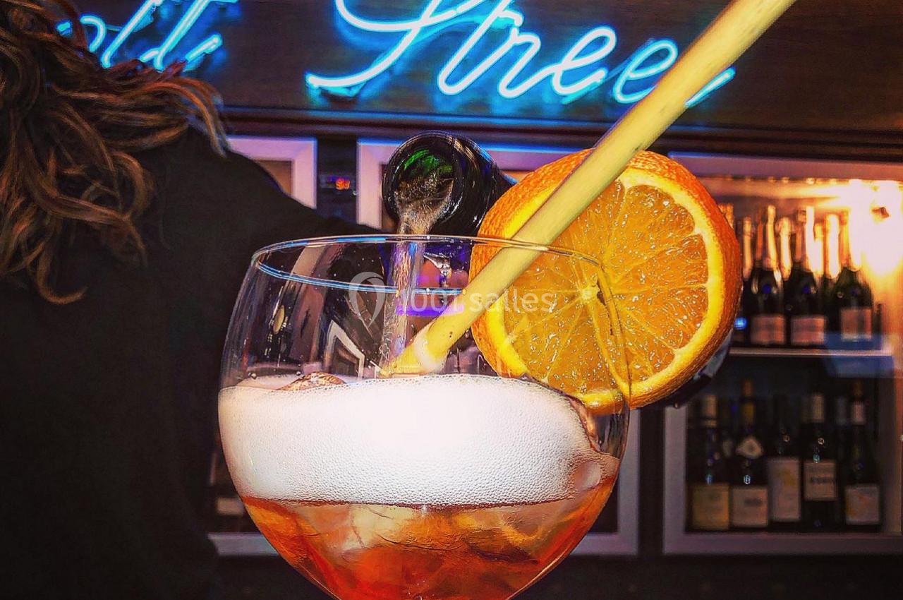 Verre de cocktail avec tranche d'orange, paille et mousse, devant un bar éclairé par une enseigne néon bleue.