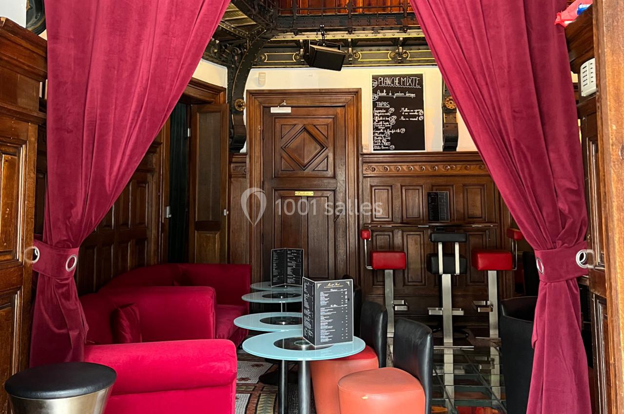 Espace intérieur avec fauteuils rouges, tables basses et rideaux ouverts, décoré de boiseries et d'un tableau noir au fond.