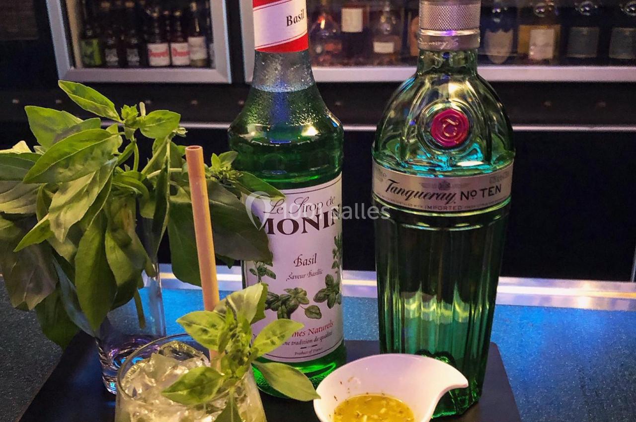 Bouteilles de sirop de basilic et de gin sur un bar, accompagnées de basilic frais, d'un cocktail et d'une sauce.