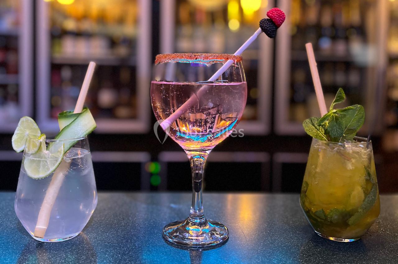 Trois cocktails colorés sur un bar, avec des garnitures de citron vert, menthe et fruits décoratifs.