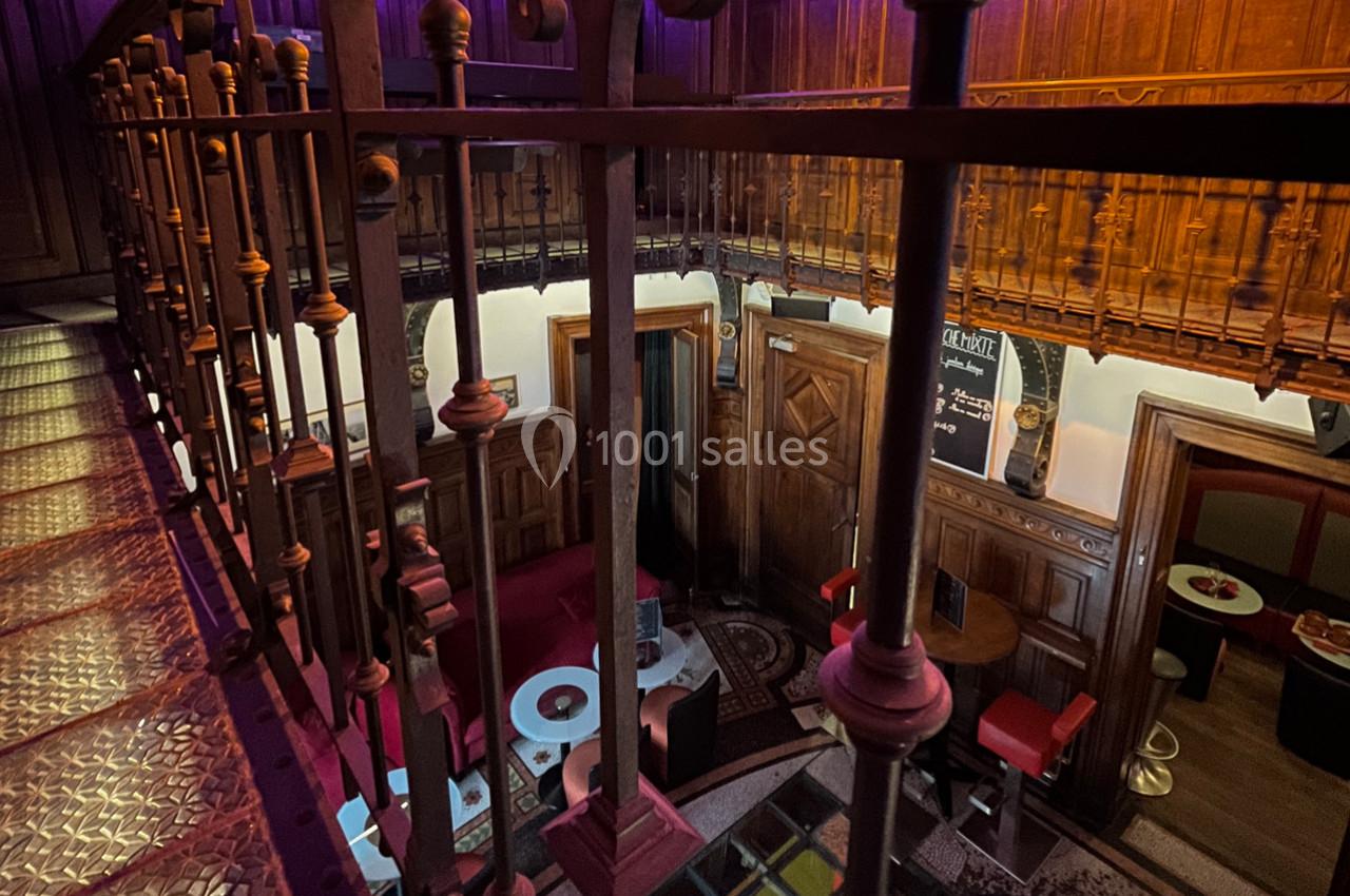 Vue intérieure d'un espace avec mezzanine en bois, balustrade métallique et coin salon avec tables et banquettes rouges.