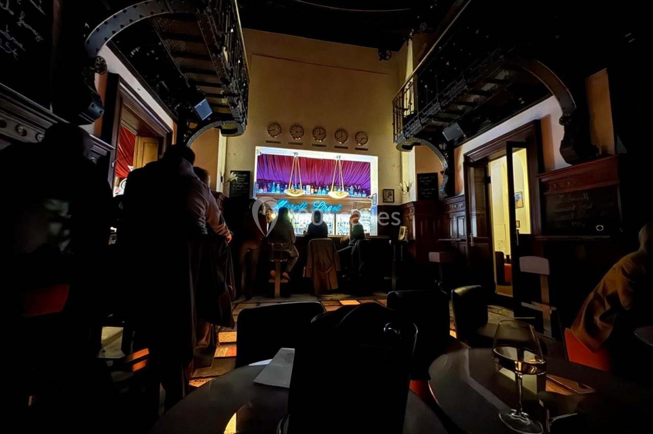 Intérieur sombre d'un bar avec des clients debout et assis, éclairé par une scène lumineuse en arrière-plan.