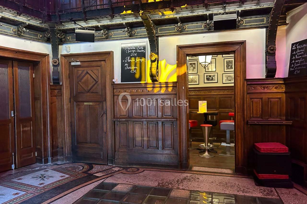 Intérieur d'un bâtiment avec boiseries, porte en bois, carrelage au sol et vue sur un espace avec tabourets rouges.