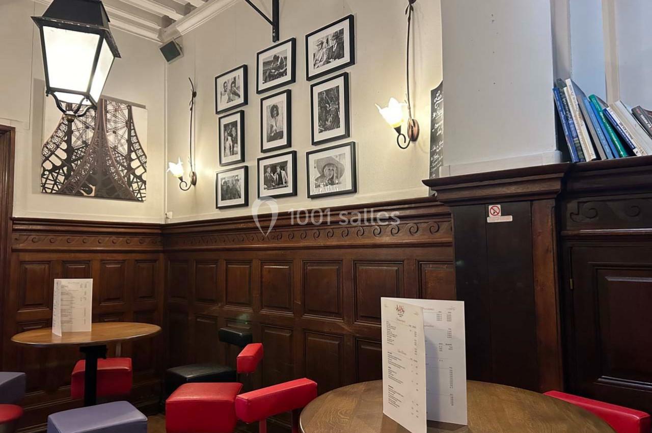 Salle de café avec murs en bois, tables rondes, chaises colorées, photos encadrées et une grande lampe suspendue.