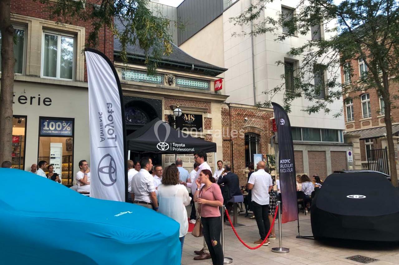 Groupe de personnes rassemblées devant un stand Toyota sur une place urbaine, entouré de bâtiments et d'arbres.