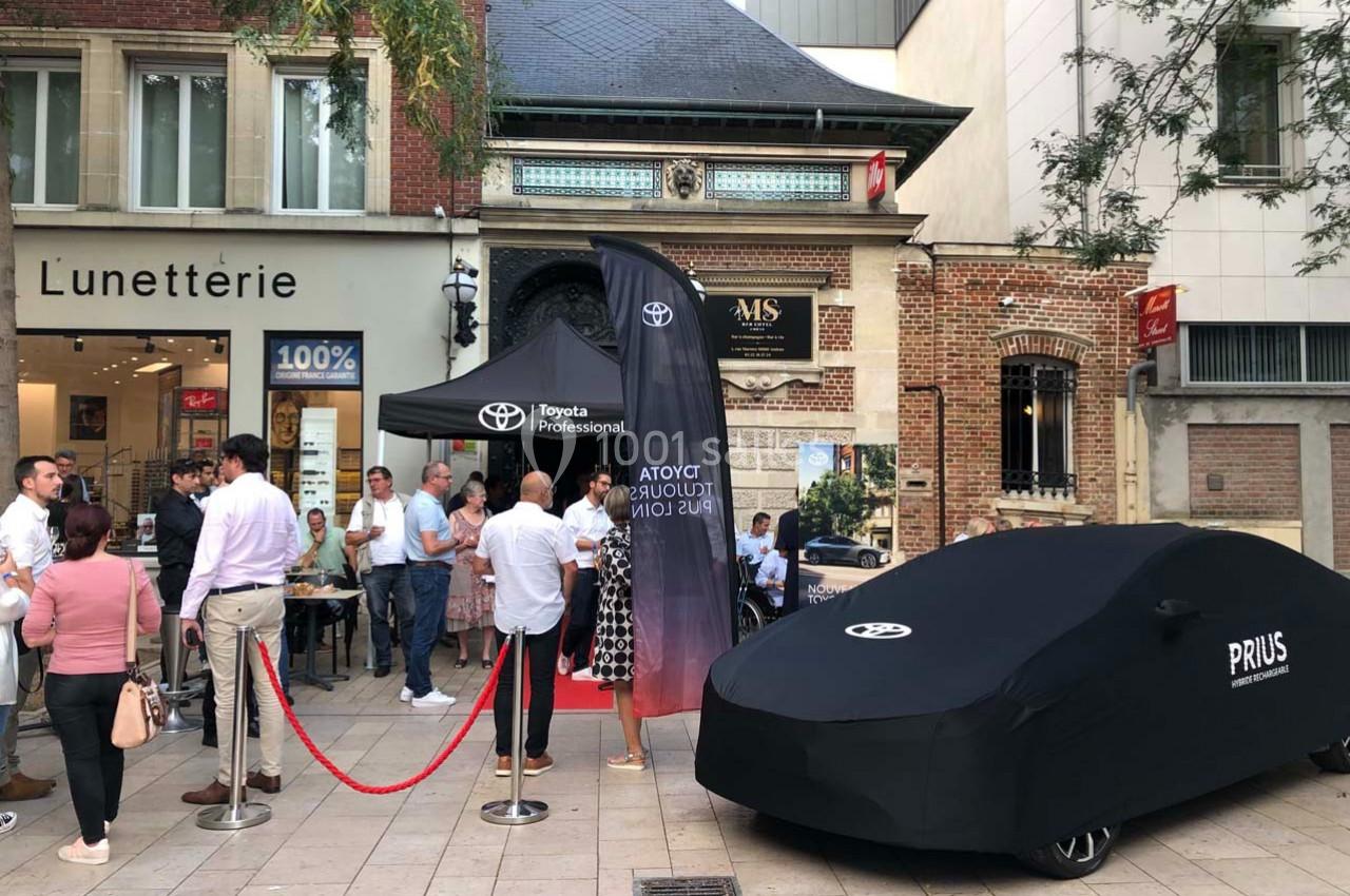 Stand Toyota avec une voiture Prius sous bâche, entouré de passants dans une rue commerçante.