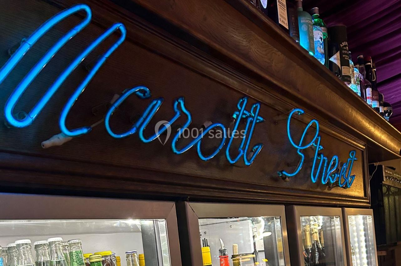 Enseigne néon bleue ’Marott Street’ sur un meuble en bois, avec des bouteilles et des boissons en arrière-plan.