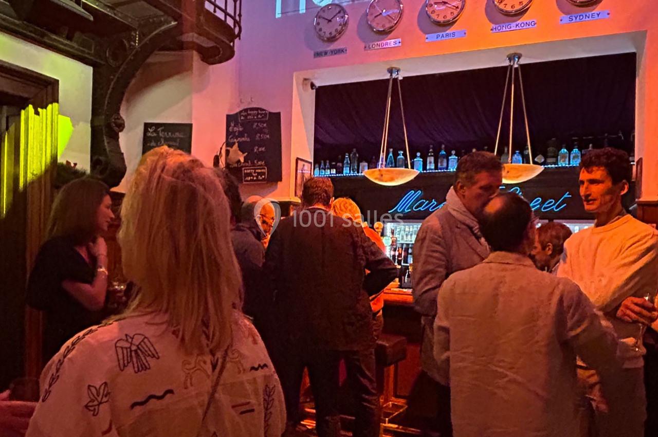 Des personnes discutent dans un bar à l'ambiance tamisée, avec une horloge murale affichant plusieurs fuseaux horaires.