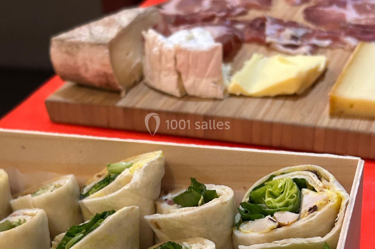 Plateau de wraps garnis de salade en premier plan, planche de charcuterie et fromages en arrière-plan flou.