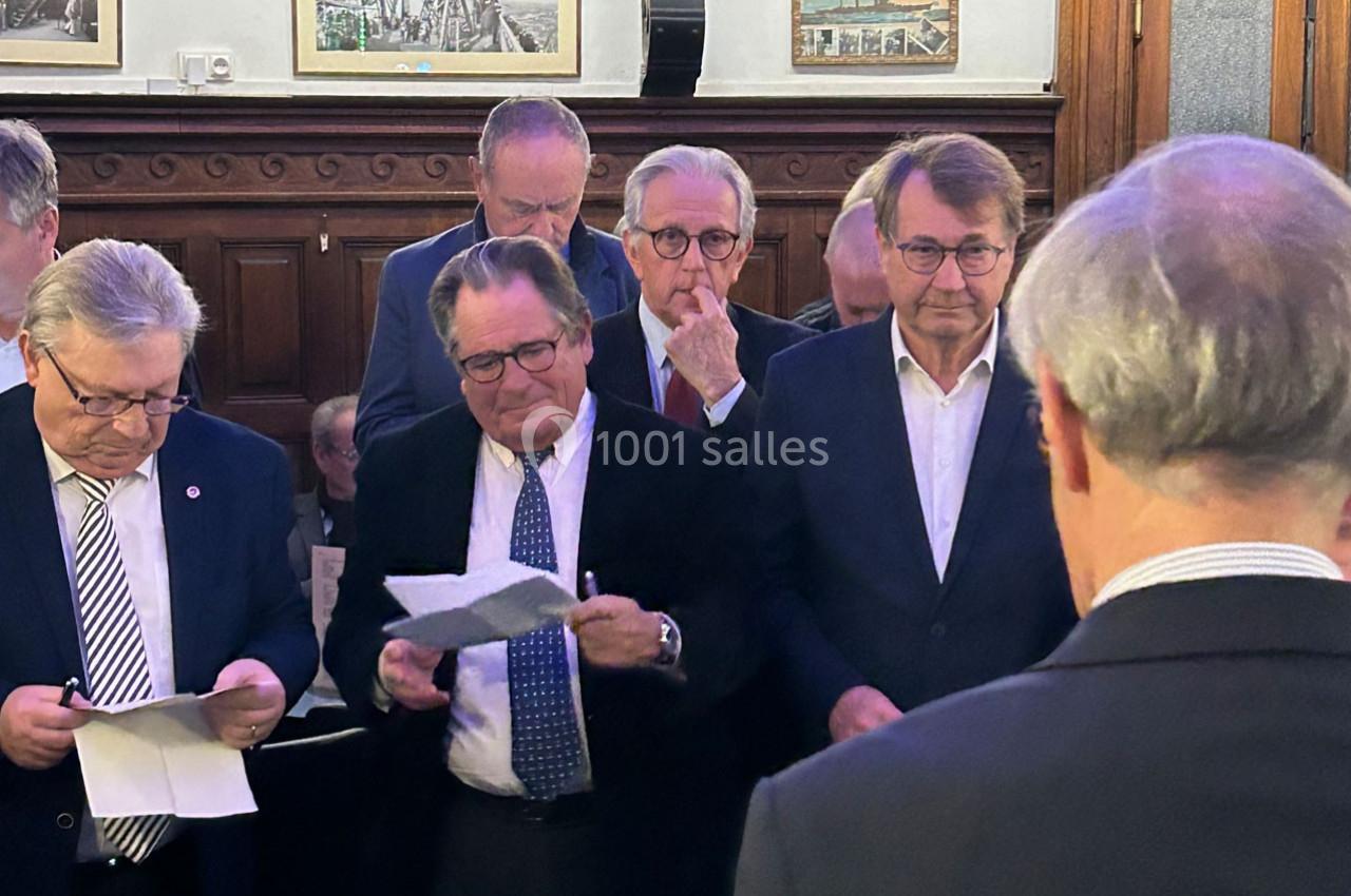 Des hommes en costume lisent des documents dans une salle avec des boiseries et des cadres accrochés au mur.