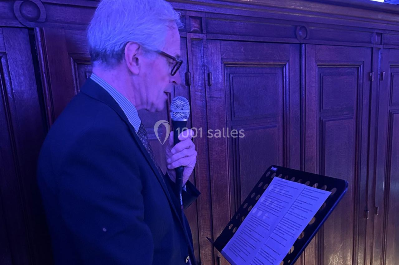Un homme âgé en costume parle dans un micro, lisant un texte sur un lutrin, devant un fond en bois sombre.