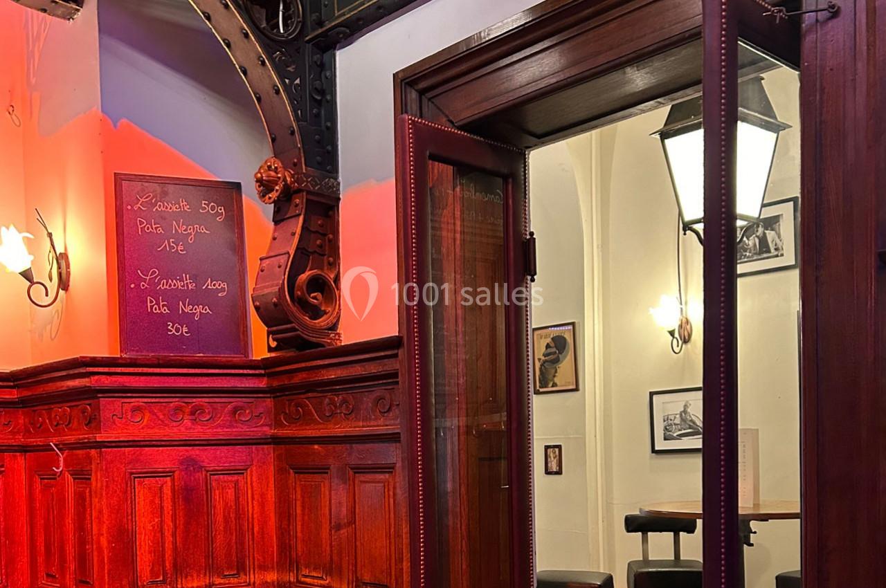 Intérieur d'un restaurant avec boiseries, tableau de menu écrit à la craie et porte vitrée donnant sur une salle éclairée.