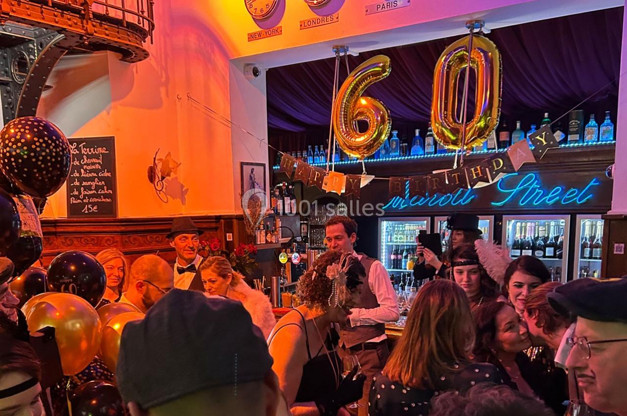 Fête d'anniversaire dans un bar décoré avec des ballons dorés ’60’, des invités en tenue rétro et une ambiance festive.