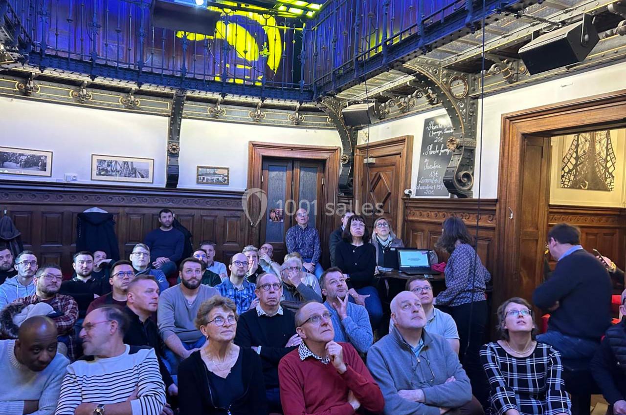 Un groupe de personnes assises dans une salle à la décoration boisée, écoutant une présentation ou un discours.