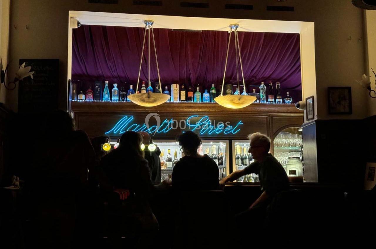 Un bar éclairé avec des bouteilles alignées, un néon ’Marott Street’ et des personnes assises au comptoir.