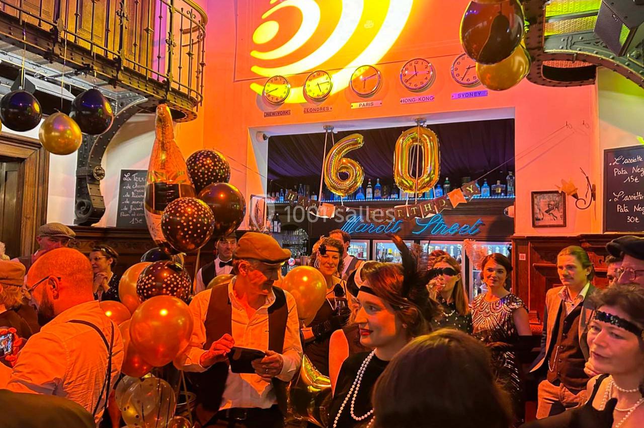 Personnes en tenue rétro célébrant un 60e anniversaire dans un bar décoré de ballons dorés et noirs.