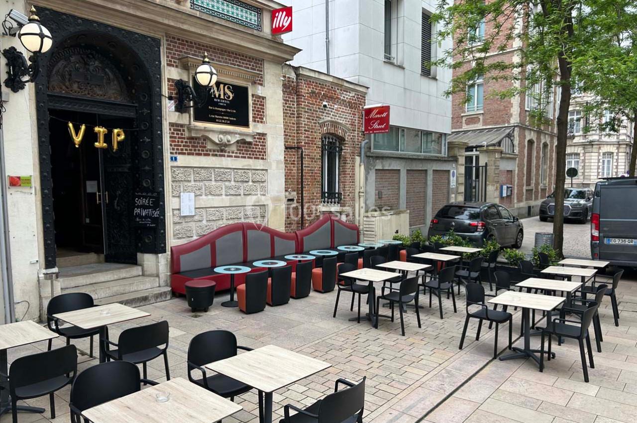Terrasse extérieure d'un café avec tables et chaises alignées, située devant une façade en briques et un bâtiment moderne.