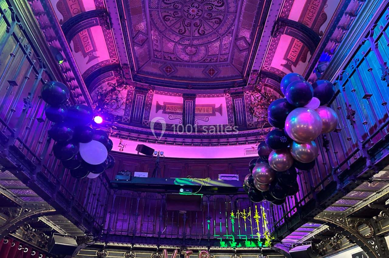 Plafond décoratif et balustrades ornées dans une salle festive avec des ballons noirs et métalliques suspendus.