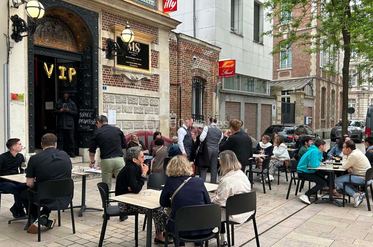 Terrasse animée d'un café avec des clients attablés, située devant une façade en briques et un panneau ’VIP’.