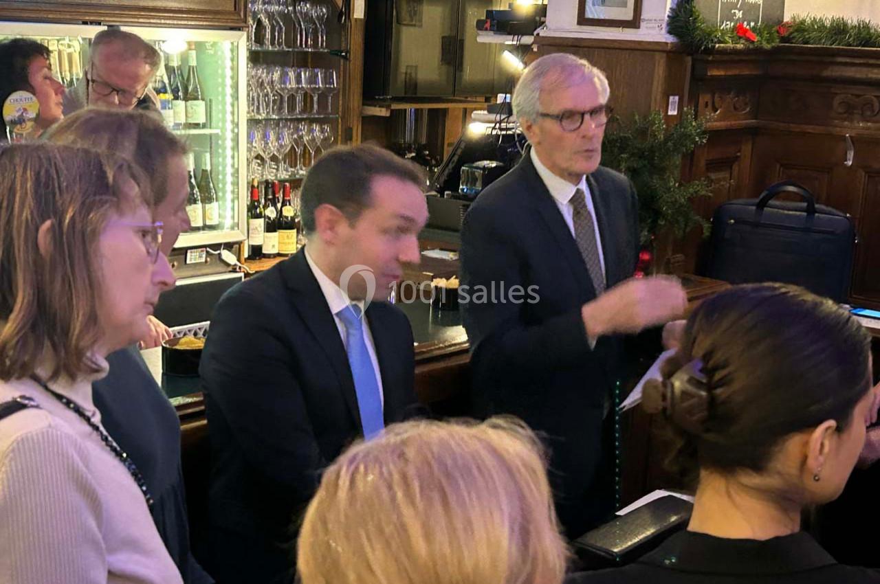 Des personnes discutent debout dans un bar ou restaurant, avec des bouteilles et des verres en arrière-plan.