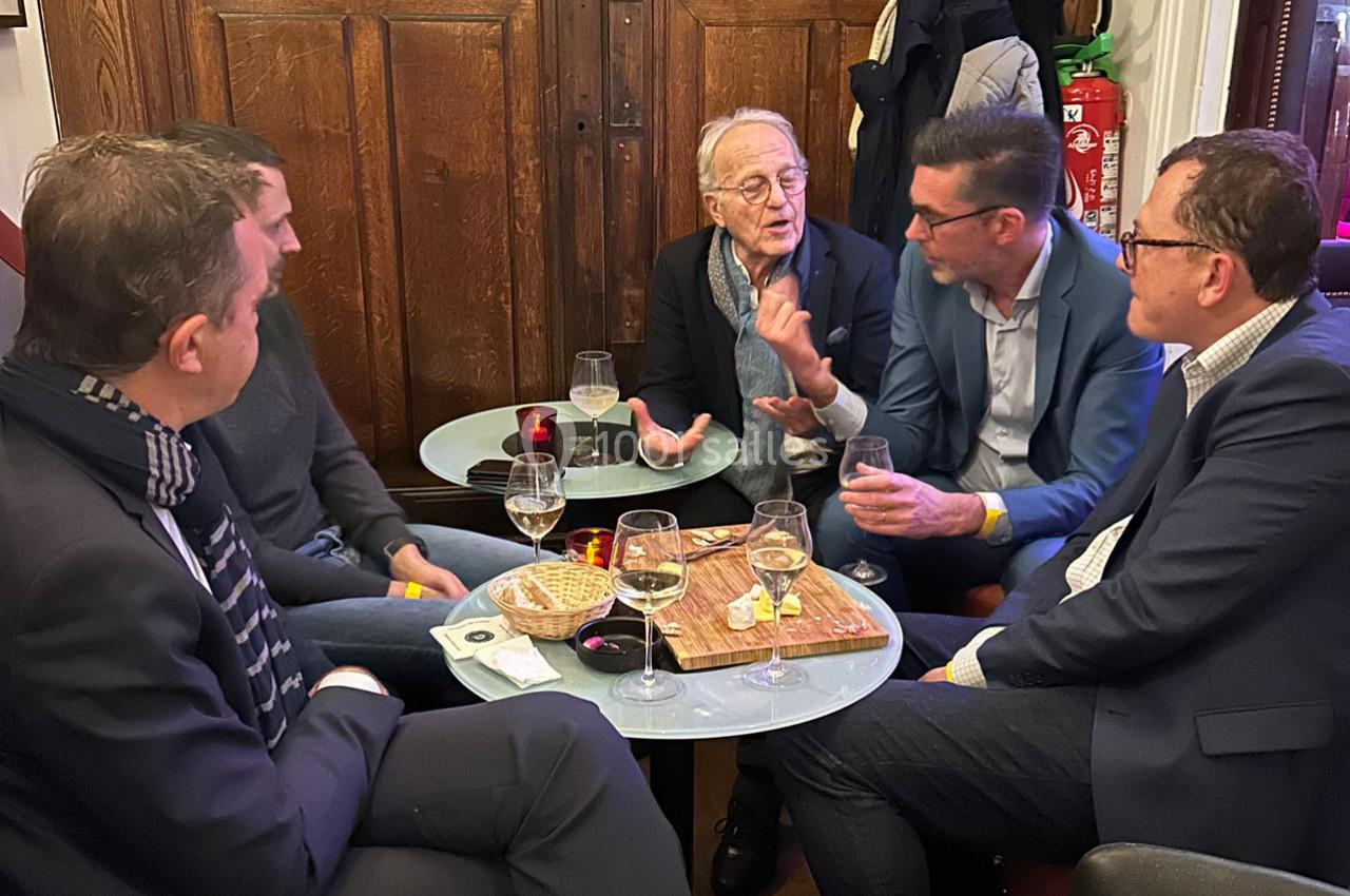 Cinq hommes discutent autour de petites tables avec des boissons et des collations dans un cadre intérieur chaleureux.