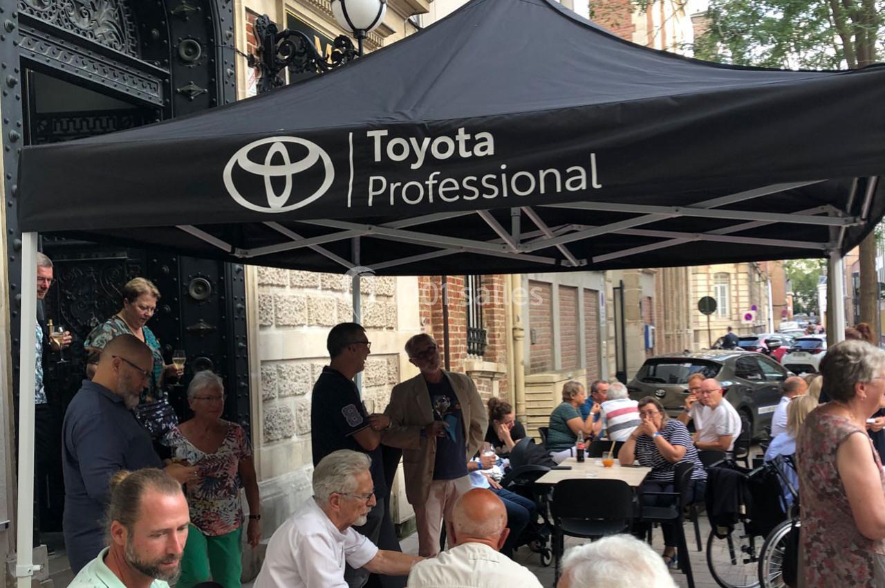 Un groupe de personnes assises et debout sous un auvent noir marqué ’Toyota Professional’ dans une rue animée.