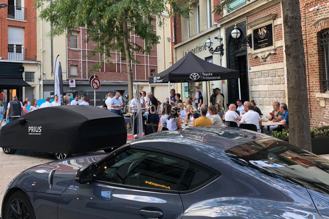Rassemblement devant un café avec des voitures exposées, dont une Toyota sous bâche et une sportive grise au premier plan.