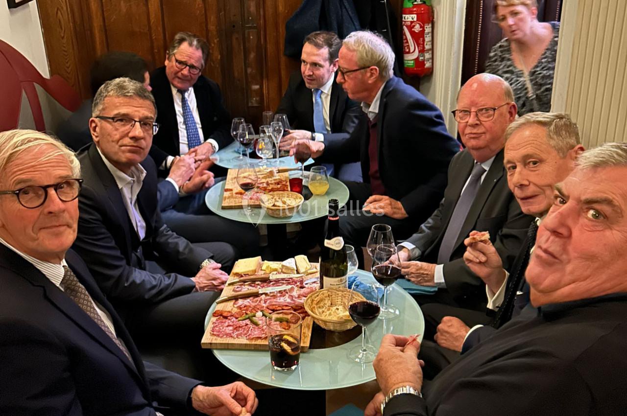 Un groupe d'hommes en costume partageant un apéritif avec charcuterie et vin autour de petites tables dans un intérieur…