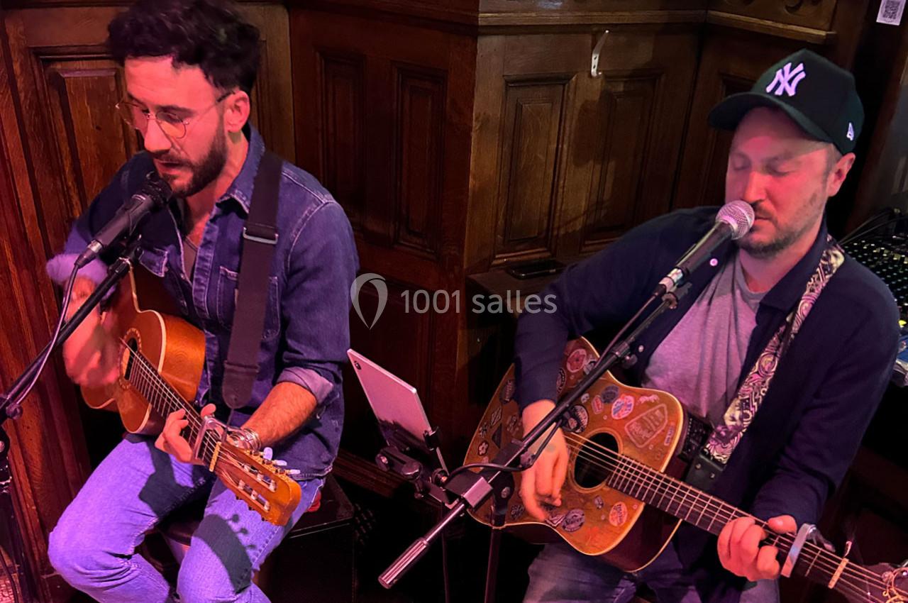 Deux musiciens jouent de la guitare et chantent dans un espace intérieur avec un décor en bois.