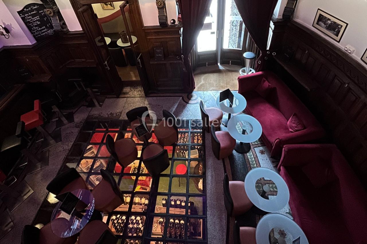 Vue plongeante d'un salon de bar avec tables, fauteuils, canapé rouge et sol en verre révélant des bouteilles en dessous.