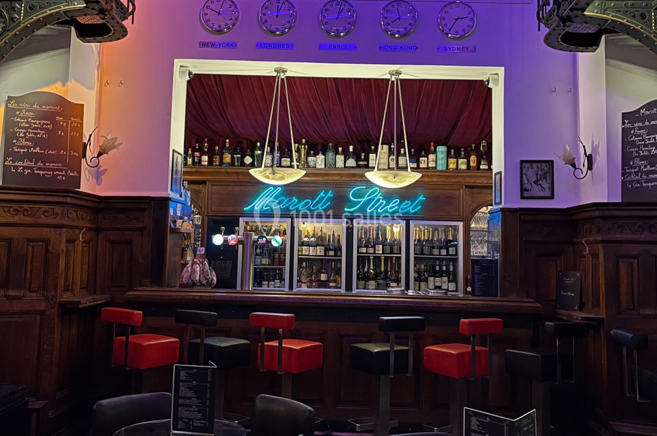 Bar avec comptoir éclairé par un néon bleu, tabourets rouges et noirs, étagères remplies de bouteilles, ambiance tamisée.