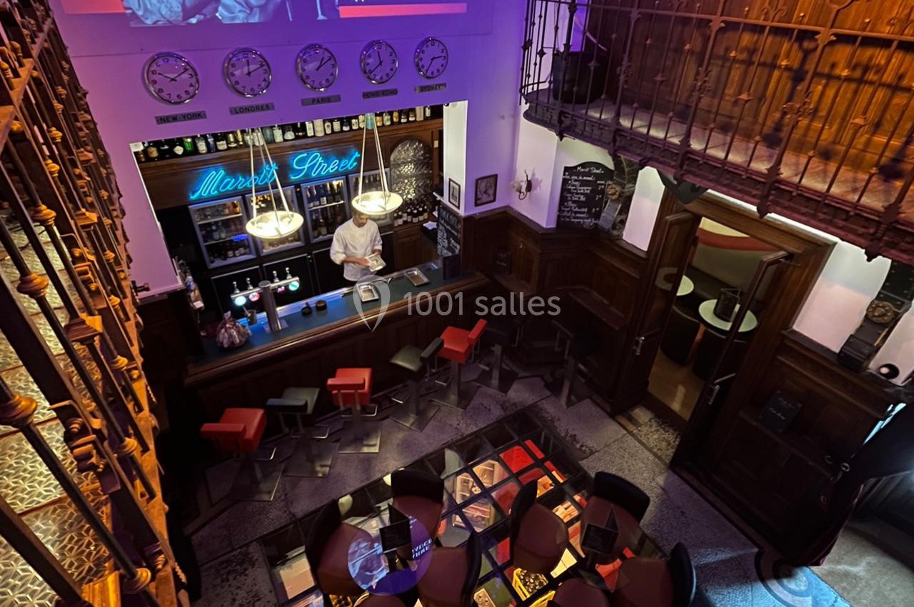 Vue plongeante d'un bar avec un comptoir éclairé, des tables colorées et une décoration intérieure en bois.