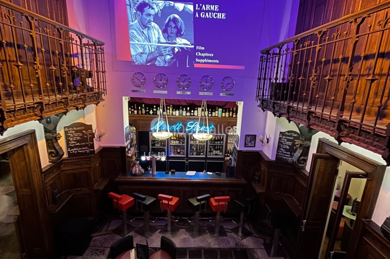 Vue d'un bar rétro avec comptoir éclairé, tabourets rouges et projection d'un film en arrière-plan.