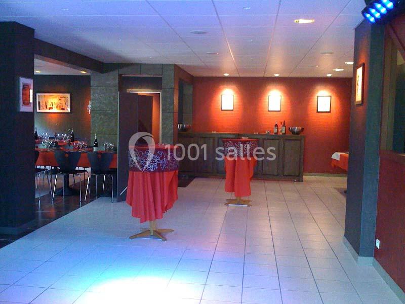 Salle de réception avec tables hautes drapées de rouge, un bar en bois et des tables dressées en arrière-plan.